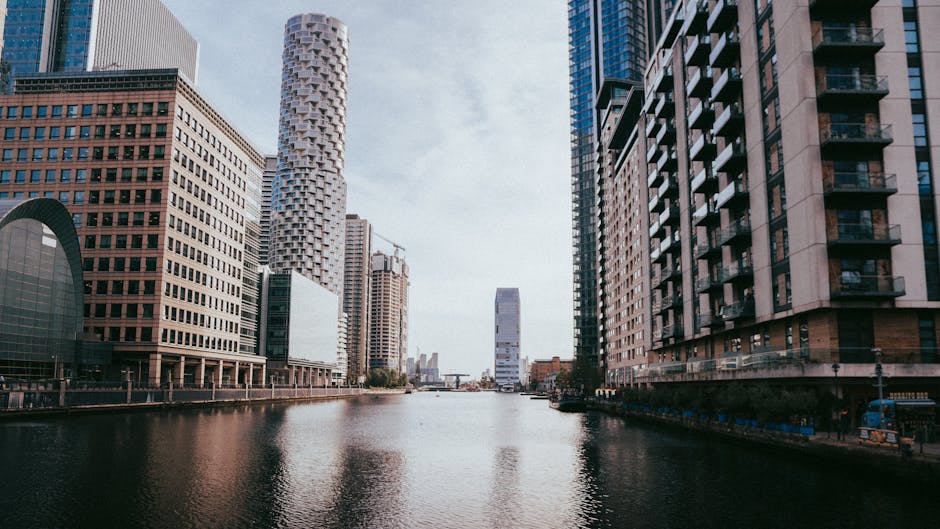 A cluster of modern high-rise buildings in an urban cityscape, with the tallest building featuring a sleek glass facade reflecting the sky. Surrounding structures include a variety of architectural st