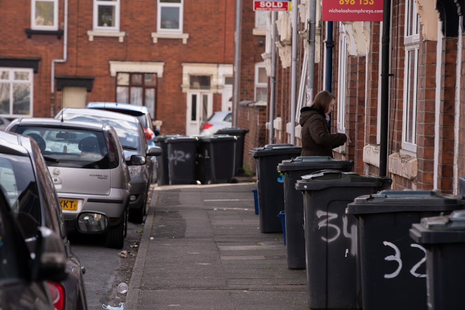 An overflowing collection of rubbish bins and scattered waste bags situated on a paved area in front of a commercial building, with a grey metal communal recycling container labeled for mixed paper an
